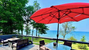 A deck with outdoor furniture, a red umbrella, and a view of the sea. - Lakefront Oak Cottage • Hot Tub & Fire Pit (Sodus)