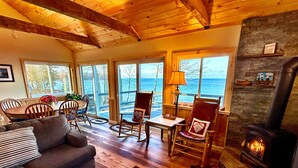A cozy living room with a fireplace, wooden furniture, and large windows overlooking a scenic view. - Lakefront Oak Cottage • Hot Tub & Fire Pit (Sodus)