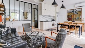 Una sala de estar moderna con un sofá de cuero negro, sillones estampados y una mesa de centro de vidrio. Un comedor con mesa y sillas de madera está adyacente a la cocina, que cuenta con gabinetes blancos y una encimera negra.