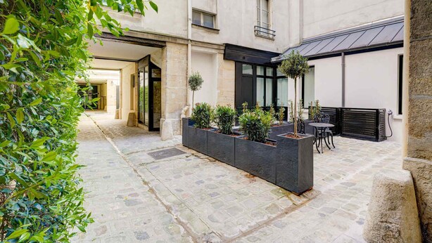 Un patio con piso de piedra, una jardinera con plantas, mobiliario exterior y un edificio de fachada oscura y ventanas.