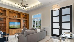 A living room with a sofa, TV, wooden shelving, and a large window. - Waterfront 4BR Marco Island home with heated pool, WiFi & kitchen. (Marco Island)
