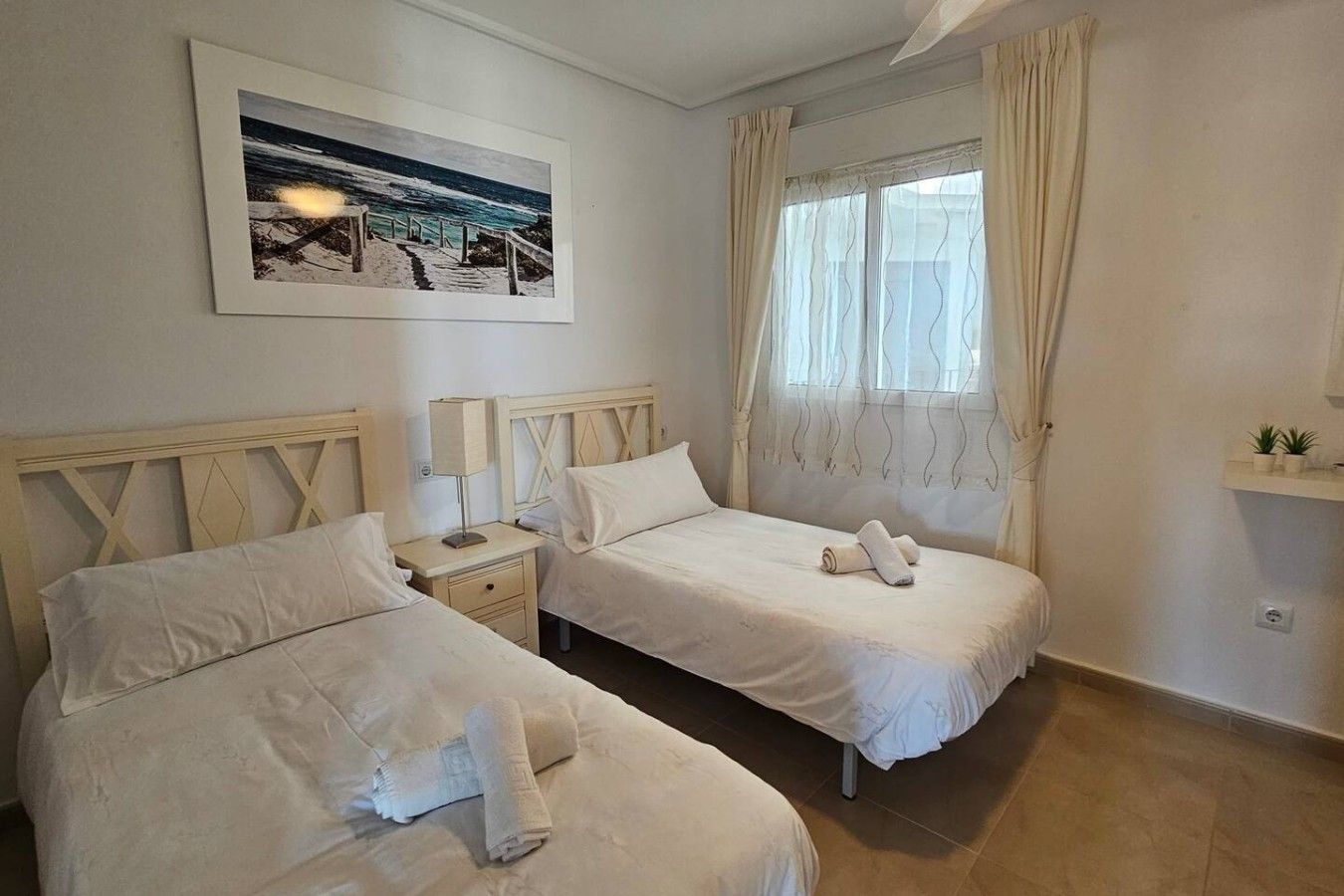 A hotel room with two beds, a nightstand, a window with curtains, a wall-mounted picture, and a small shelf with plants.