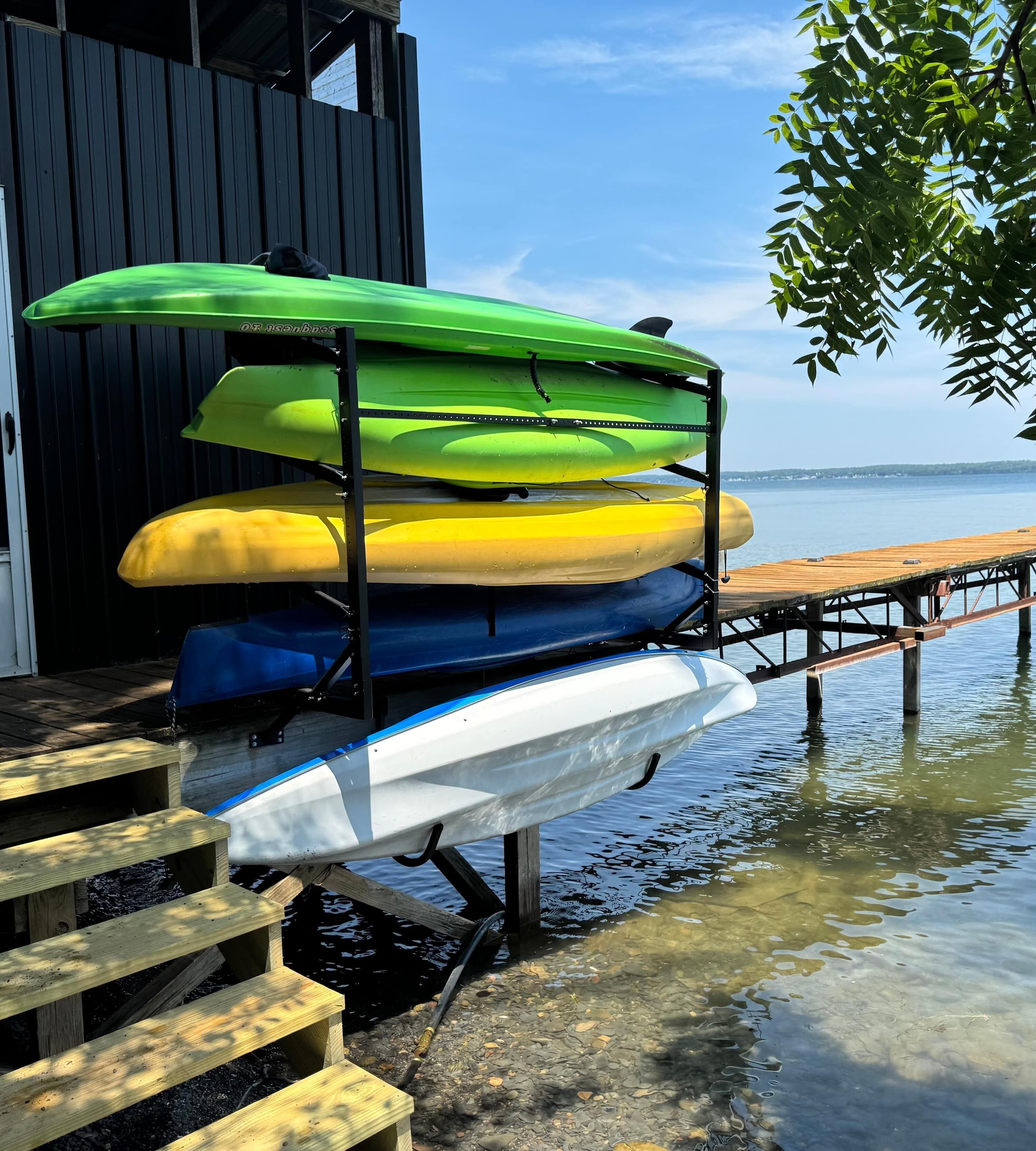 A kayak rack with multiple kayaks, a wooden dock extending into a body of water, and a building with a door in the background.
