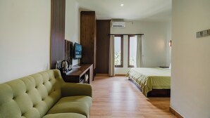 A bedroom with a bed, a sofa, a TV, and a wooden floor. - You Suite Seminyak (Legian)