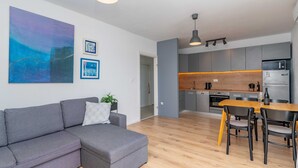 A modern living room with a grey sectional sofa, a wooden dining table with chairs, a kitchen with grey cabinets, and a large abstract painting on the wall.