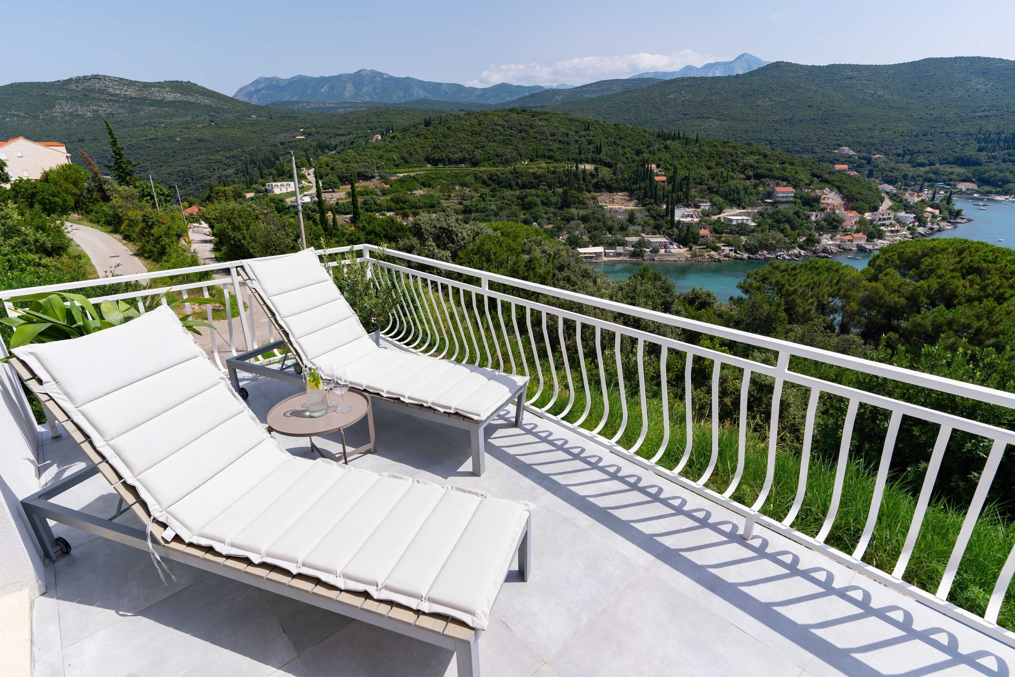 Un balcon doté de chaises longues blanches avec des coussins et d’une petite table ronde, offrant une vue panoramique sur un paysage magnifique, avec des montagnes et une étendue d’eau.
