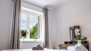 Ein ordentlich bezogenes Bett mit weißer Decke und zwei gerollten Handtüchern, ein Fenster mit Vorhängen, eine Vase mit Blumen und eine Holzkommode mit Spiegel und Orchideenpflanze.