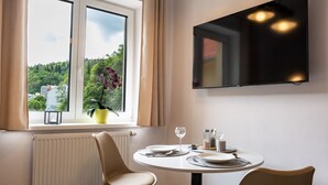 Ein Essbereich mit einem Tisch für zwei Personen, einem Fenster mit Blick auf Grünflächen und einem an der Wand montierten Flachbildfernseher.
