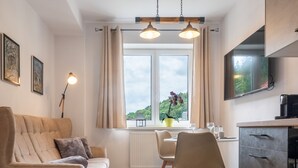 Ein modernes Wohnzimmer mit Sofa, Esstisch mit Stühlen, Küchenzeile mit Schränken und einem großen Fenster mit Blick auf Grünflächen.