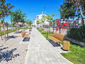 Stroll through this urban park, featuring a playground, benches, and a trash bin, surrounded by trees and buildings.