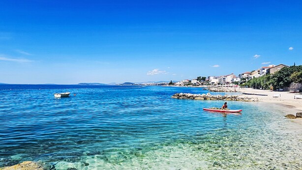 A coastal scene with clear blue waters, a rocky shoreline, and a person paddling a red kayak.