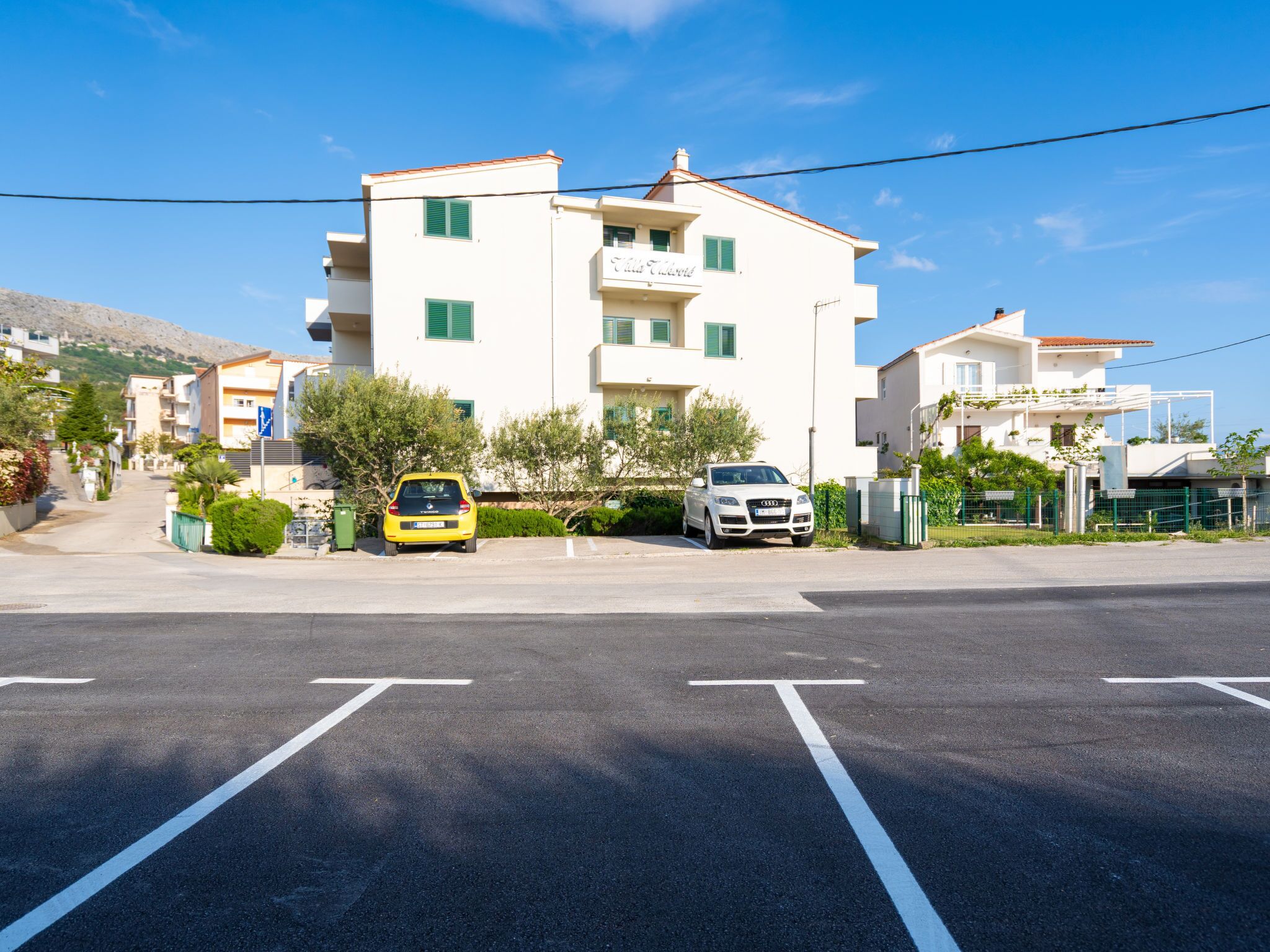 A multi-story building with a white facade and green shutters, a yellow car parked in front, and a white car in the driveway.