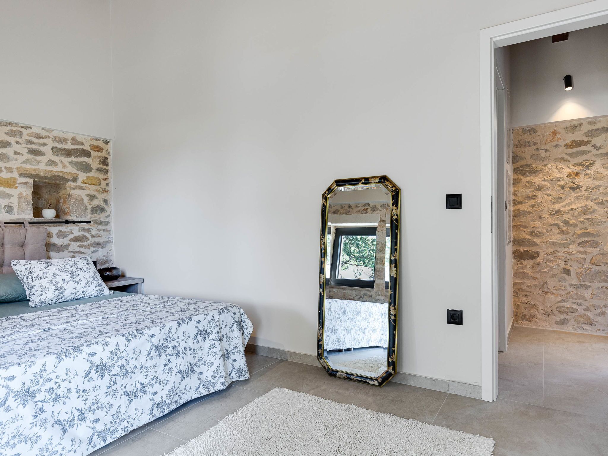 A bedroom with a stone wall, a bed with floral bedding, a large mirror, and a small shelf with decorative items.