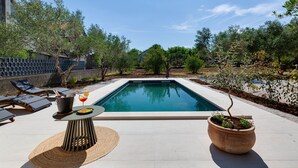 Una piscina rectangular con sillas de descanso y una mesa con bebidas, rodeada de árboles y un muro de piedra.