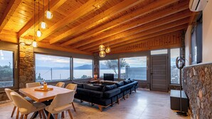 A spacious room with a wooden ceiling, stone walls, a large window, a black sofa, a dining table with chairs, and a TV. - Epitome Villa Aegina (Aegina)