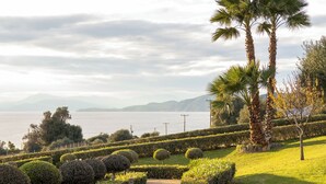 A paved pathway leading to a landscaped area with neatly trimmed bushes and palm trees, overlooking a body of water with distant mountains. - Epitome Villa Aegina (Aegina)