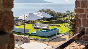 A view of a terrace with outdoor furniture, a table, and umbrellas, overlooking a body of water and mountains. - Epitome Villa Aegina (Aegina)