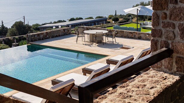 A pool with lounge chairs, a stone wall, and a view of the sea and mountains. - Epitome Villa Aegina (Aegina)