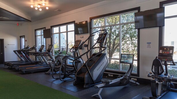 Une salle de sport équipée de tapis de course, de vélos elliptiques et de vélos d’appartement, de grandes fenêtres donnant sur la verdure et un panneau d’affichage sur le mur.