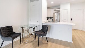 Une cuisine moderne avec des meubles blancs, un réfrigérateur en acier inoxydable et une table à manger en verre, accompagnée de deux chaises.