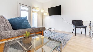 Un salon moderne comprenant un canapé, une table basse, un fauteuil, un téléviseur fixé au mur et une fenêtre avec des stores.
