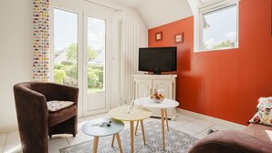 Ein Wohnzimmer mit einem braunen Sessel, einem runden Tisch mit Blumenvase, einem Fernseher auf einem weißen Schrank und einem großen Fenster mit Blick auf Grünflächen.