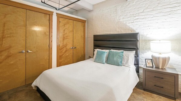 Un dormitorio con una cama, dos armarios de madera, una mesita de noche con lámpara y una pared de ladrillo blanco.