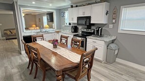A dining area with a wooden table and chairs, a kitchen with white cabinets and appliances, and a trash can.
