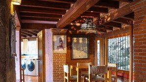 A rustic interior with exposed wooden beams, a stone floor, a dining area with wooden furniture, and a view through large windows.