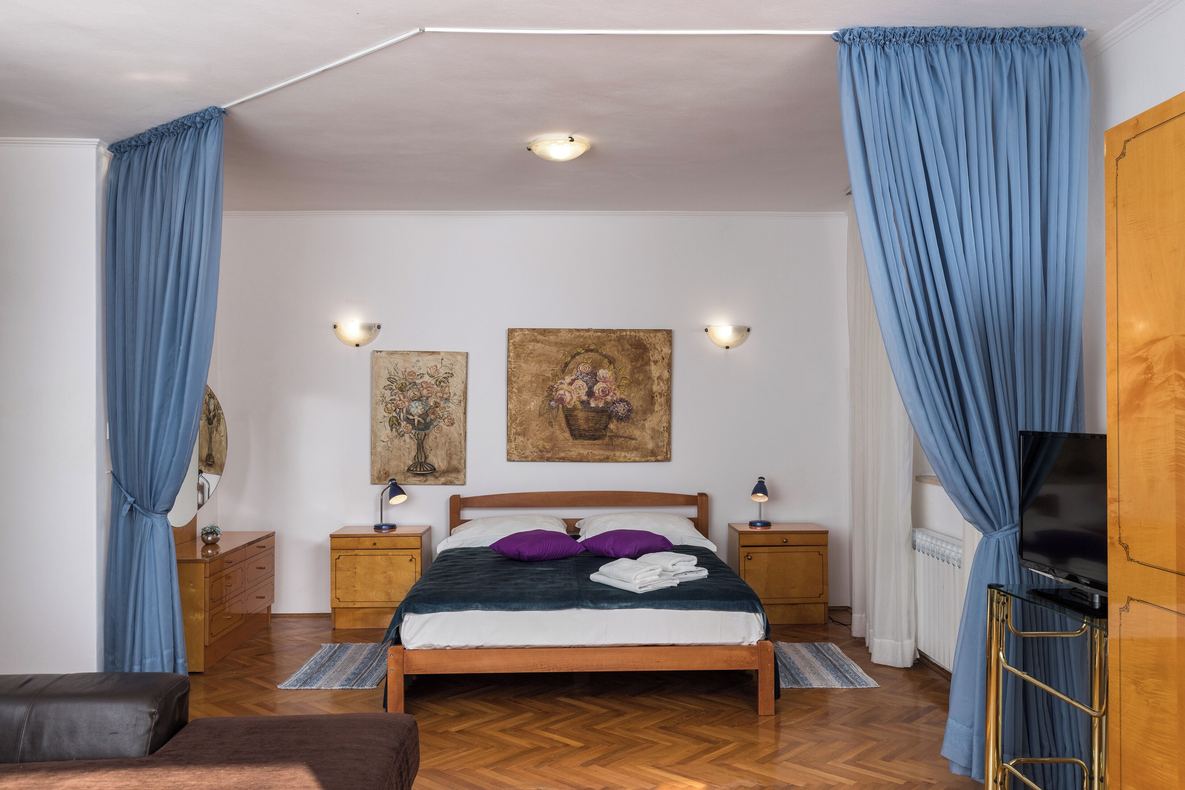 A bedroom with a wooden bed, blue curtains, two bedside tables with lamps, a TV, and artwork on the wall.