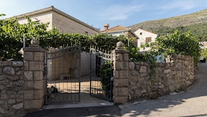 A stone-walled property with a gated entrance, a stone building with a tiled roof, and a hillside in the background. - Apartment Vukic (Dubrovnik)