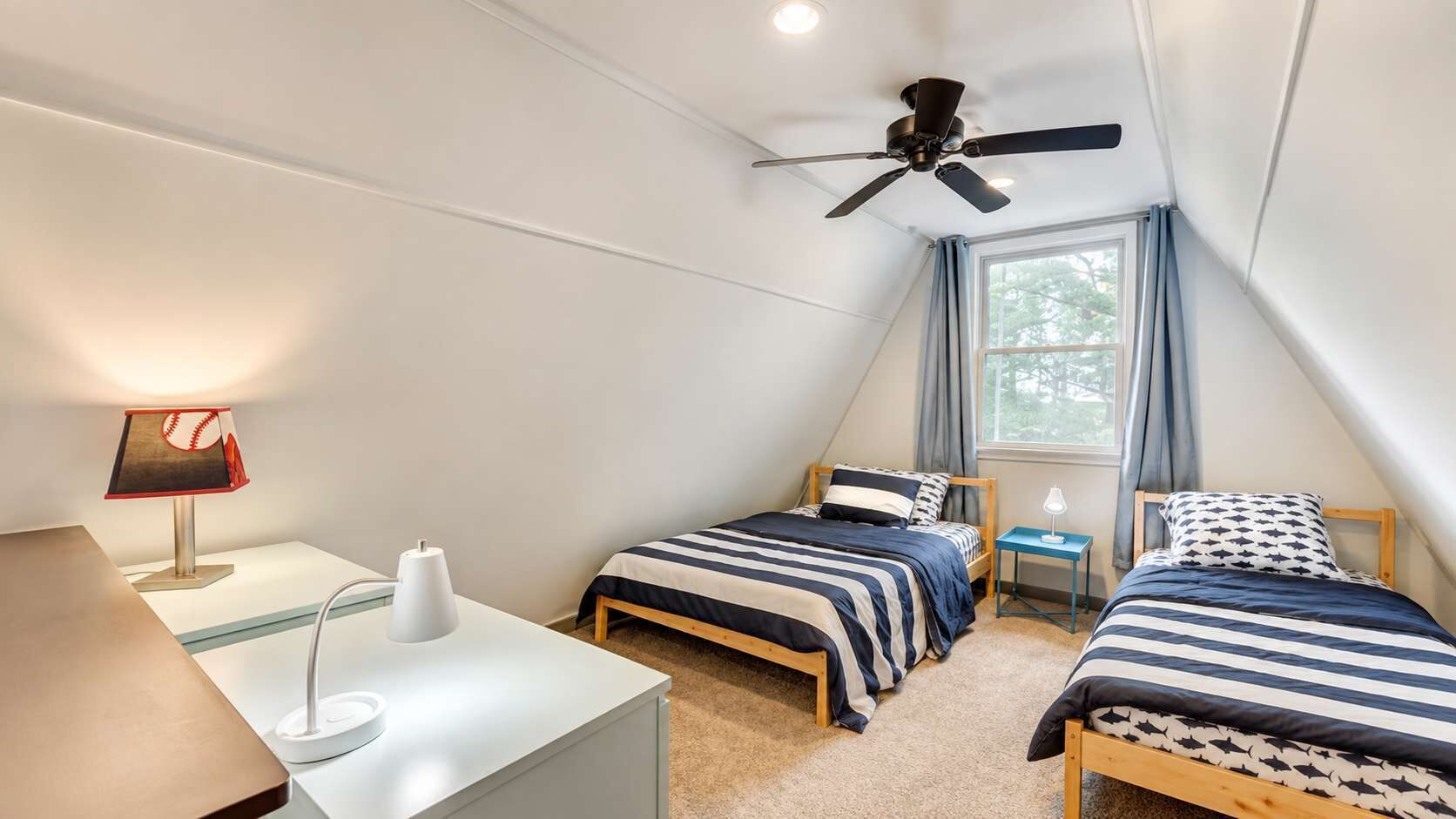 Quarto com duas camas, uma escrivaninha, abajur, ventilador de teto e janela com cortinas.