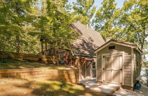 Un rustico chalet con facciata in legno, un garage e un piccolo deposito, circondato da alberi e un pontile in legno.