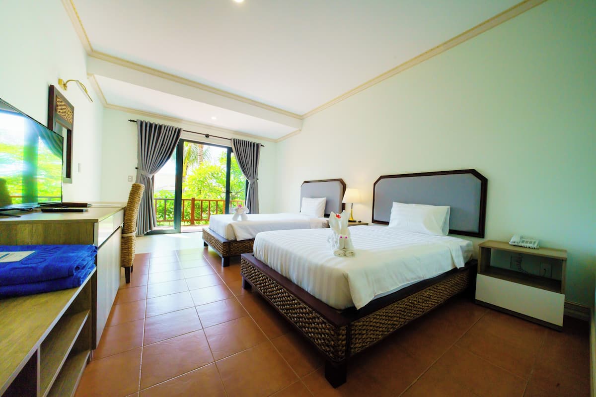 A hotel room with two beds, a TV, a desk, and a balcony with curtains.