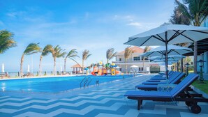 A pool area with lounge chairs and umbrellas, palm trees, and a building in the background. - Blooming Lotus Resort (Tien Thanh)