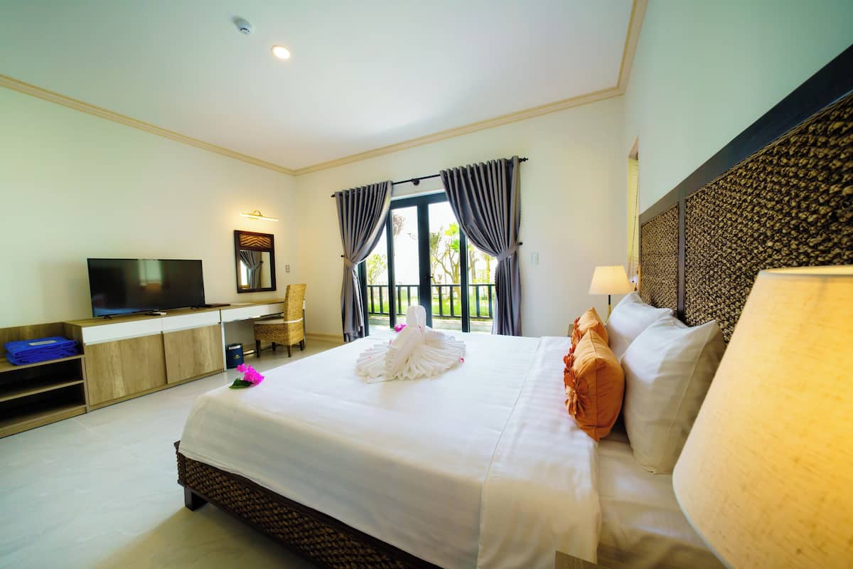 A hotel room with a large bed, a flat-screen TV, a desk with a chair, and a view of the outdoors through large windows.