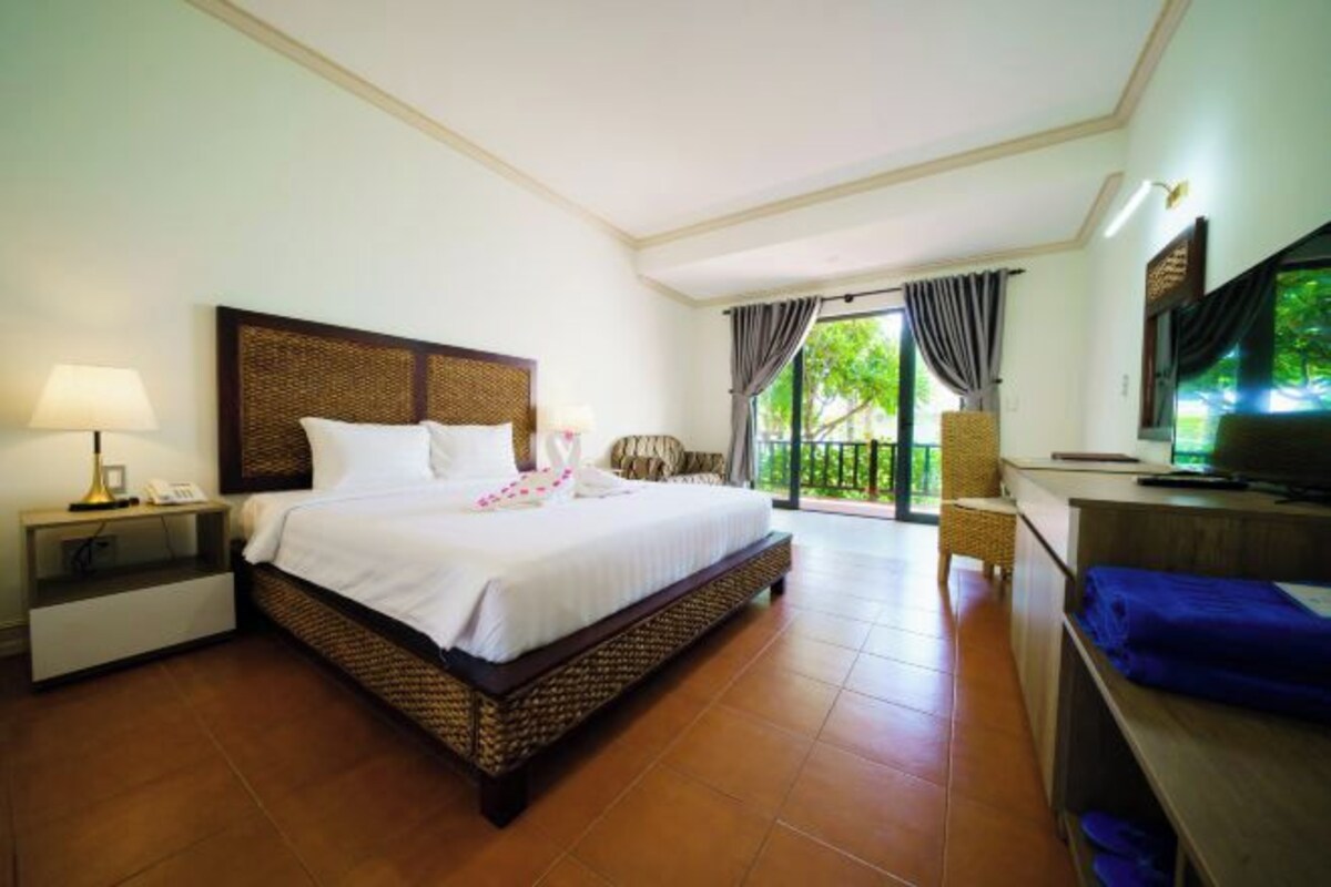A hotel room with a large bed, a desk with a TV, a chair, and a balcony with curtains.