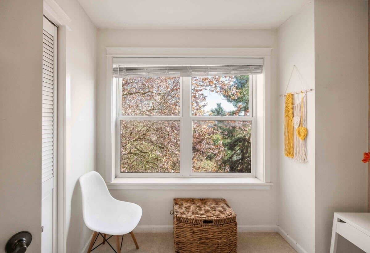 A room with a large window, a white chair, a wicker basket, and a wall-mounted macrame piece.