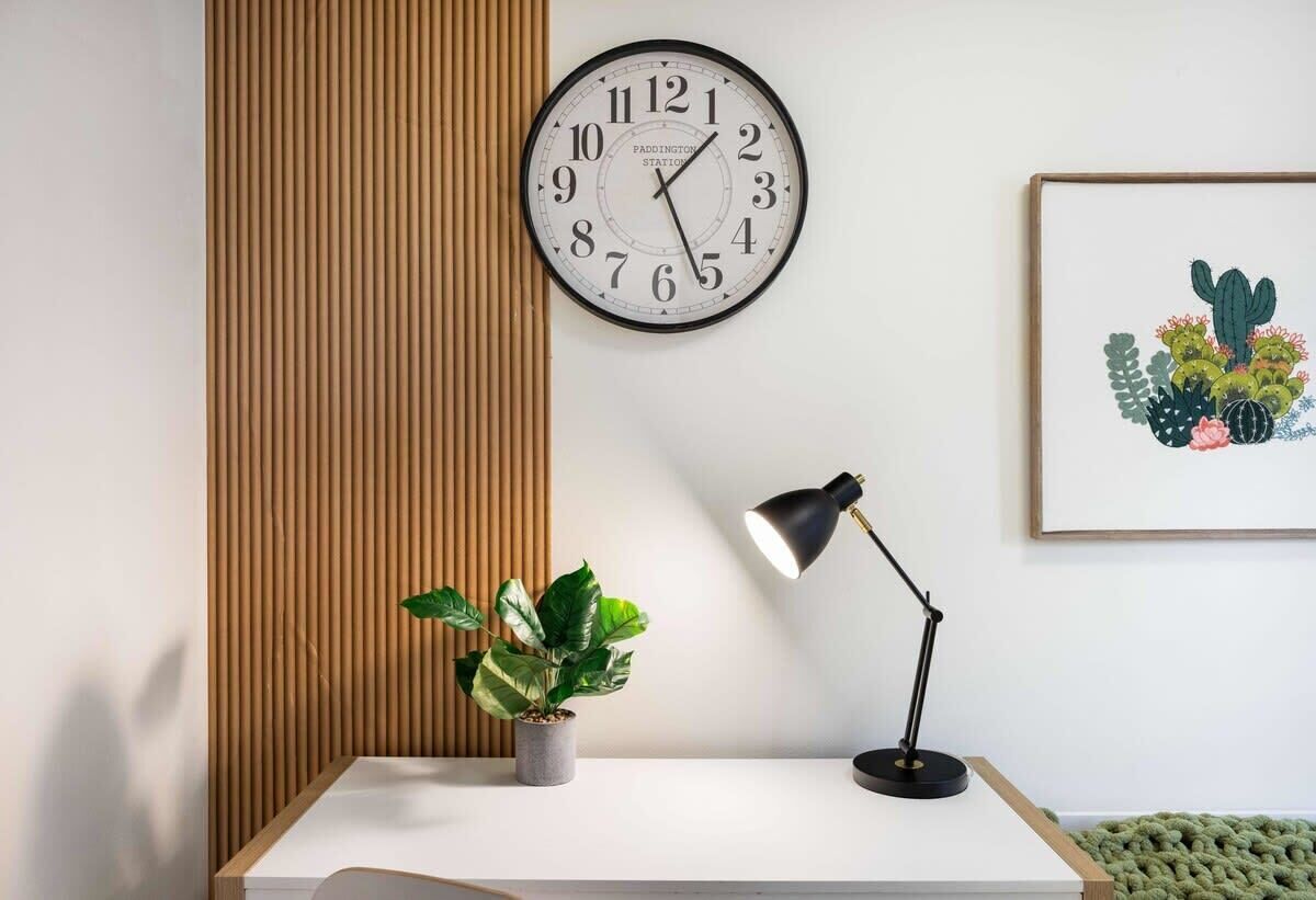 A wall-mounted clock, a desk with a plant, a desk lamp, and a framed picture of cacti.