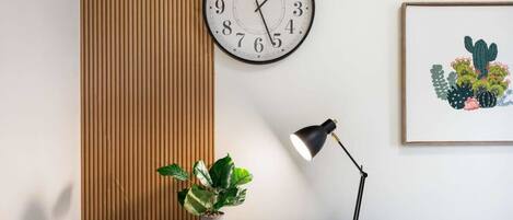 A wall-mounted clock, a desk with a plant, a desk lamp, and a framed picture of cacti.