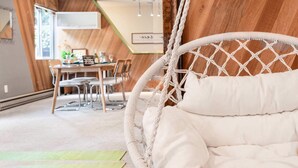 Ein Zimmer mit Holzwand, einem Hängekorbstuhl, einem Essbereich mit Tisch und Stühlen sowie Blick ins Freie.