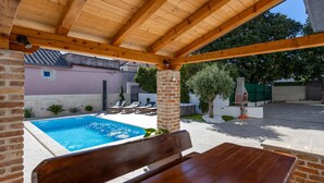 A covered outdoor area with a pool, lounge chairs, and a brick column.