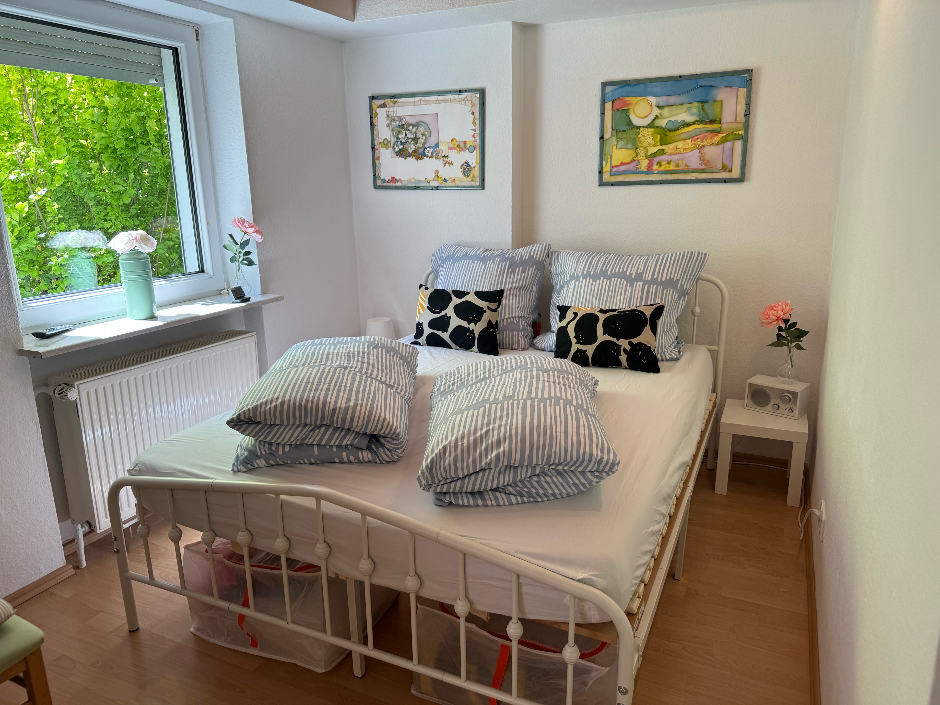 A neatly arranged bedroom with a bed, pillows, a nightstand, and a window with a view of greenery.