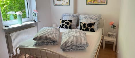 Un dormitorio ordenado con una cama, almohadas, una mesita de noche y una ventana con vistas al verdor.