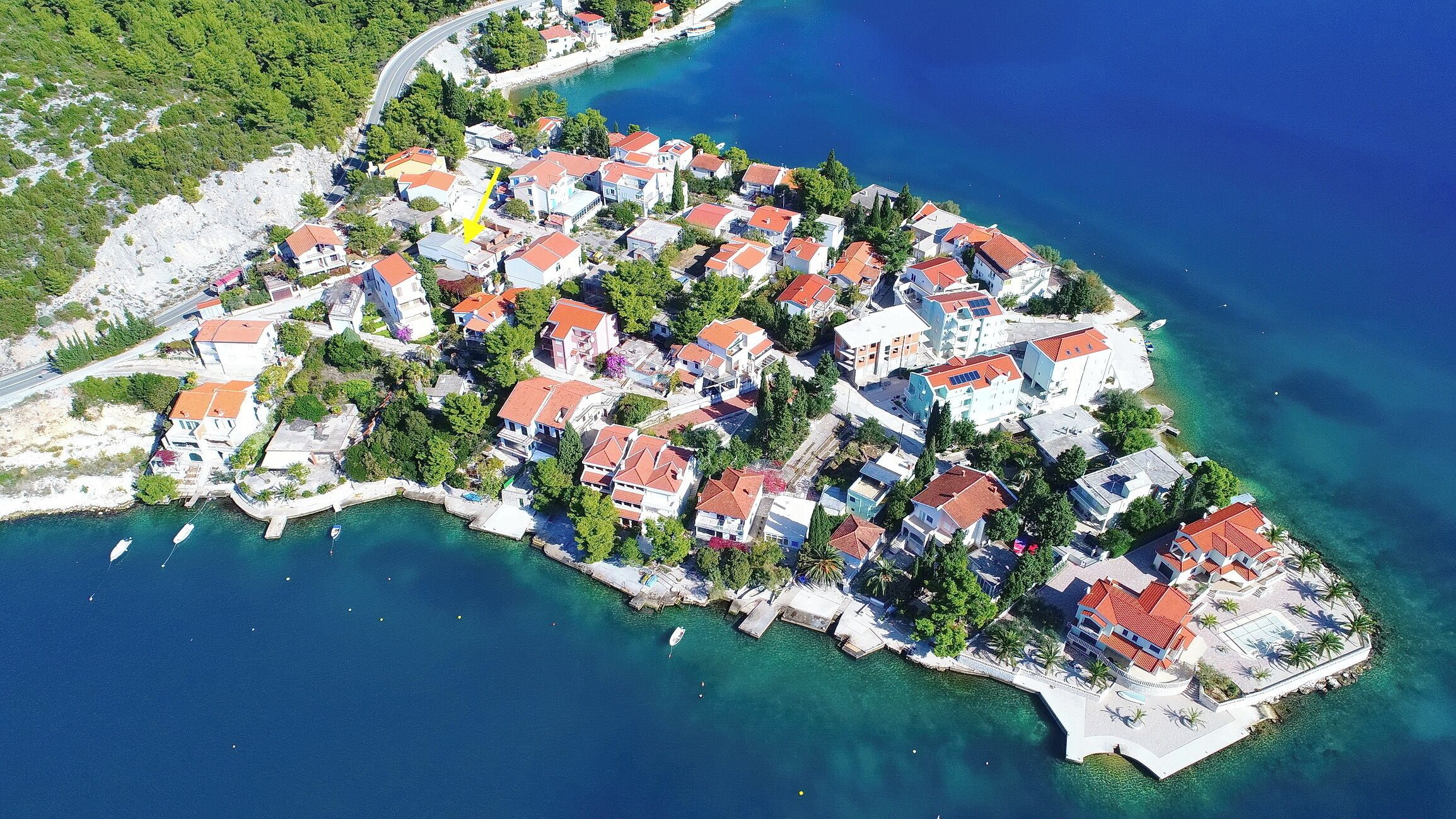 An aerial view of a coastal village with houses, clear blue waters, and a road curving through the area.