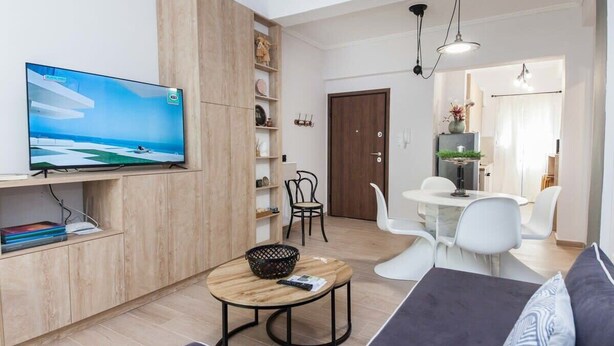Ein modernes Wohnzimmer mit Flachbildfernseher, Holzschrank, rundem Couchtisch, einer Couch und einem Essbereich mit weißen Stühlen und einem kleinen Tisch.