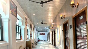 A corridor with ceiling fans, wall-mounted lamps, and potted plants. - Hotel Ambassador  (Varanasi)
