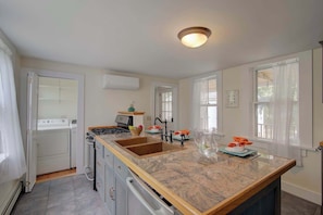 A modern kitchen with a central island, a built-in oven, and a stove. There are two windows with curtains, a wall-mounted air conditioner, and a ceiling light fixture. - Short drive to town and popular beaches! (Orleans)