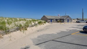 En byggnad vid stranden med en blå dörr, en parkeringsplats och en sanddyn med växtlighet.