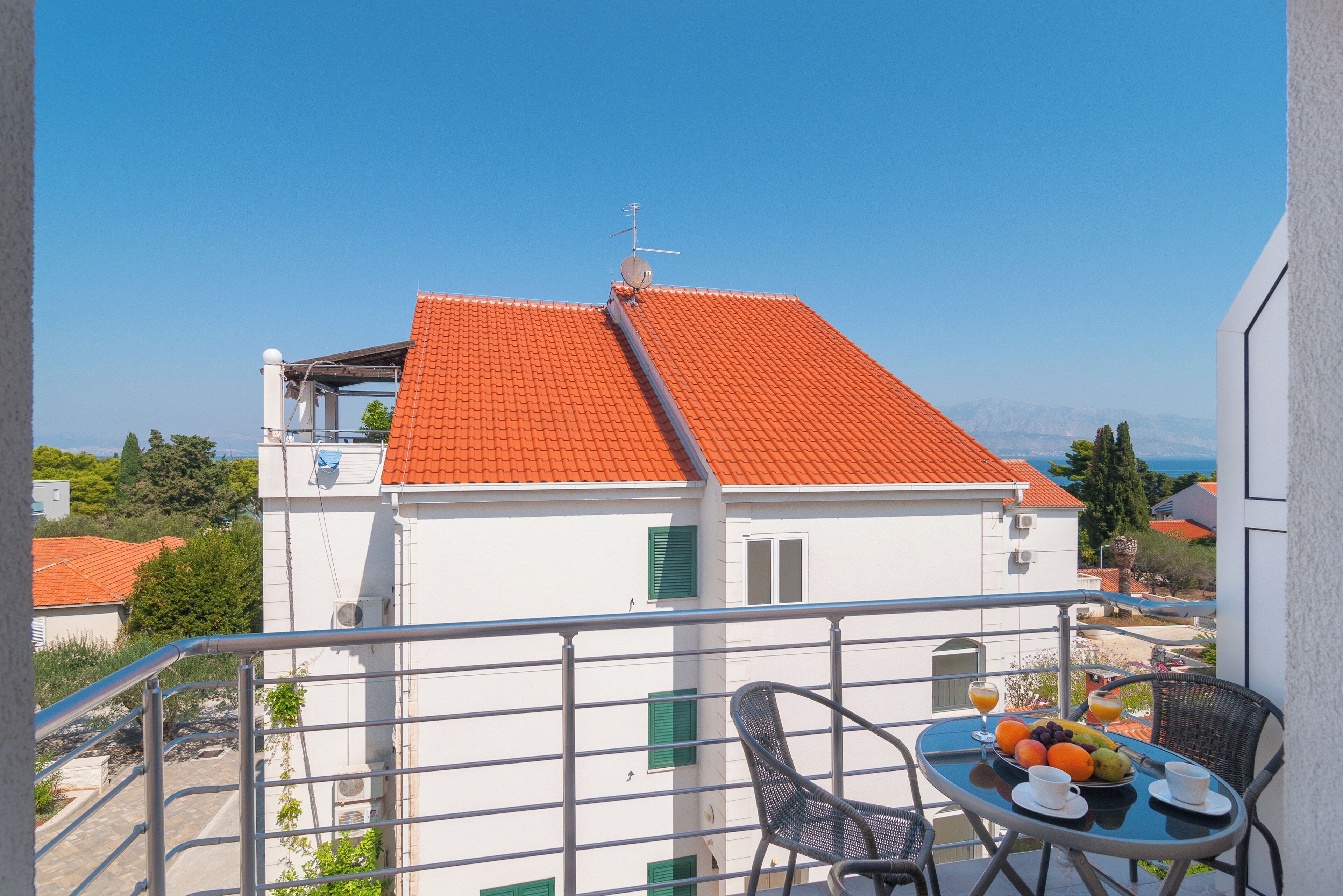 A balcony with a table set for breakfast, a red-tiled roof, and a view of the sea and mountains.